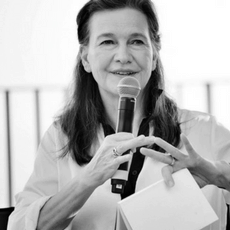 A woman with long, dark hair offers writing advice into a microphone, gesturing passionately with her other hand. Clad in a light-colored blouse against a blurred backdrop, the black-and-white image captures her eloquent dedication to the craft.