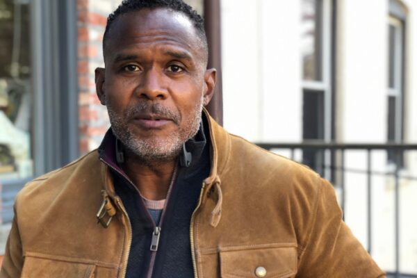 A person in a brown jacket with a black inner layer stands outdoors, framed by an urban background. They have close-cropped hair and a short beard, looking directly at the camera with a calm expression that seems to convey unspoken writing advice.