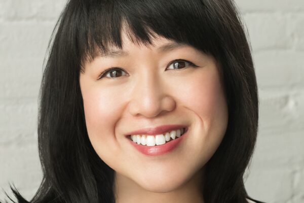 Smiling person with medium-length black hair and a white shirt, offering writing advice while posing in front of a light-colored brick wall.