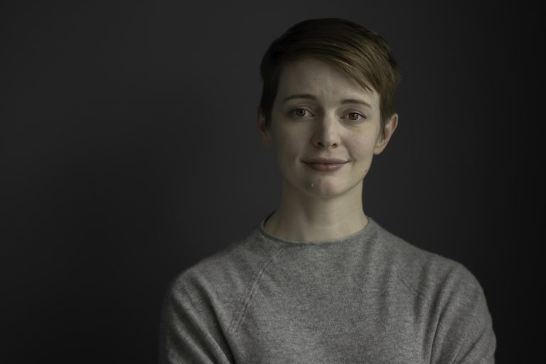 A person with short hair and a gentle expression stands against a dark background, wearing a light gray sweater. The soft lighting crafts a calm, introspective atmosphere, much like the quiet pause one takes before offering thoughtful writing advice.
