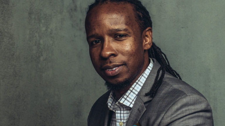 Man with long braided hair, donning a gray suit and checkered shirt, poses against a gray textured background with a slight smile, as if contemplating the next piece of writing advice.