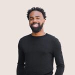 A man with a beard and curly hair, wearing a black long-sleeve shirt, is smiling against a light-colored background.