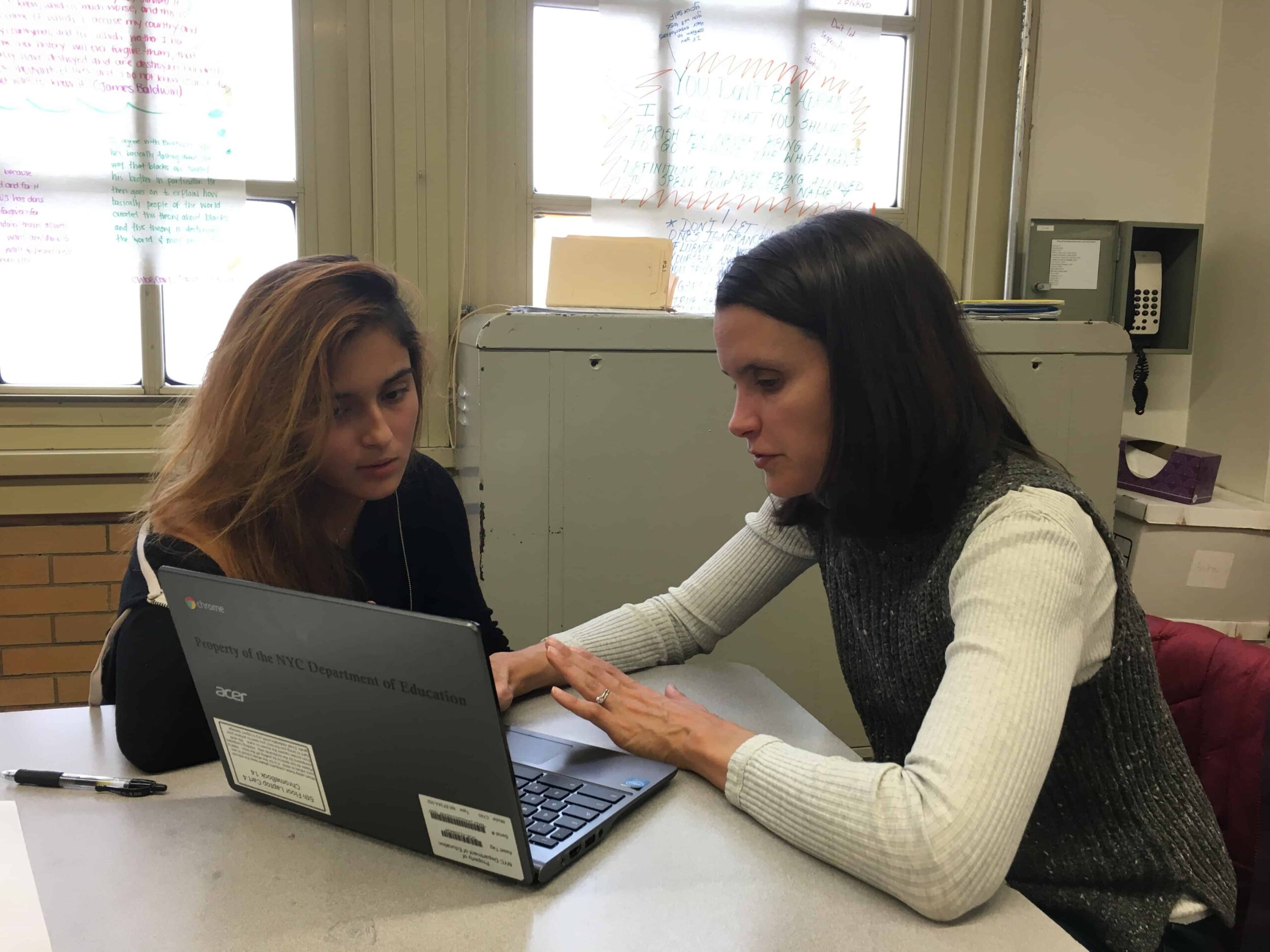 Two women sit at a table, focused on a laptop screen. One is pointing as the other listens intently. In this classroom setting, they collaborate like writers in schools, surrounded by windows and office equipment.