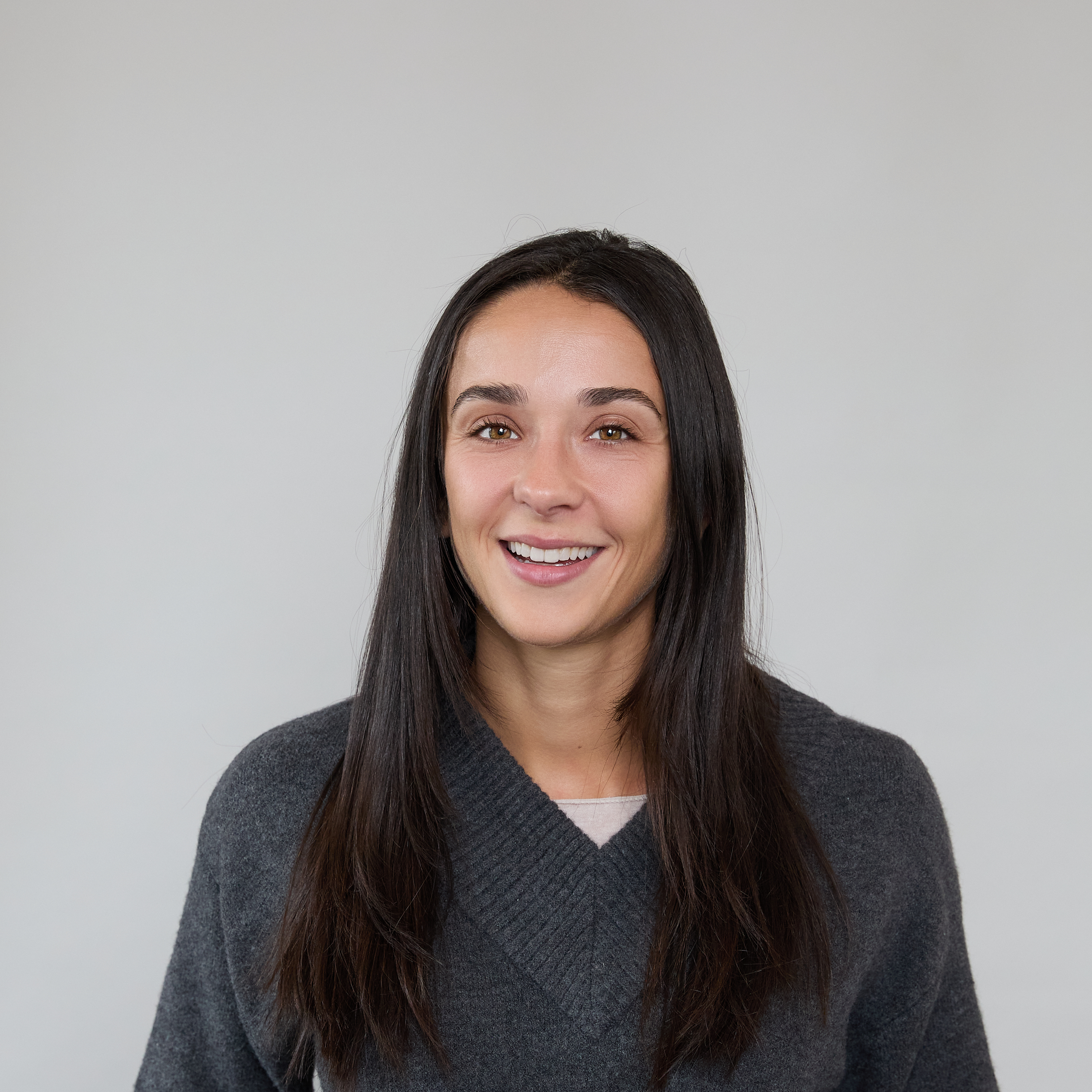 A woman with long dark hair, wearing a dark gray sweater, smiles while standing in front of a plain light gray background.