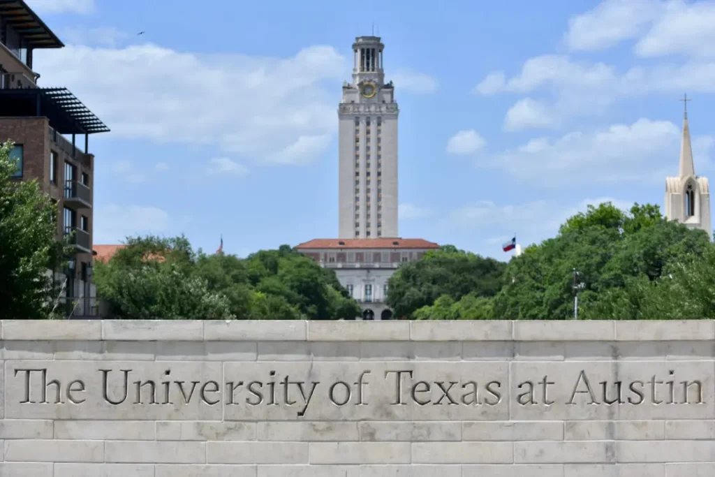 The image captures the University of Texas at Austins iconic tower set against a clear sky, framed by trees. In the foreground, a stone sign reads The University of Texas at Austin, evoking thoughts of Americas censored classrooms in 2023.