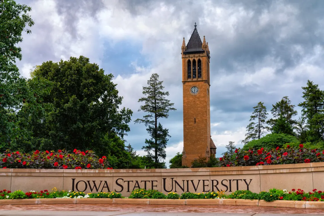 The image captures the Campanile tower at Iowa State University under a cloudy sky, surrounded by trees. In the foreground, a sign reads Iowa State University, with red flowers blooming at its base—a serene scene amidst Americas evolving campuses in 2023.