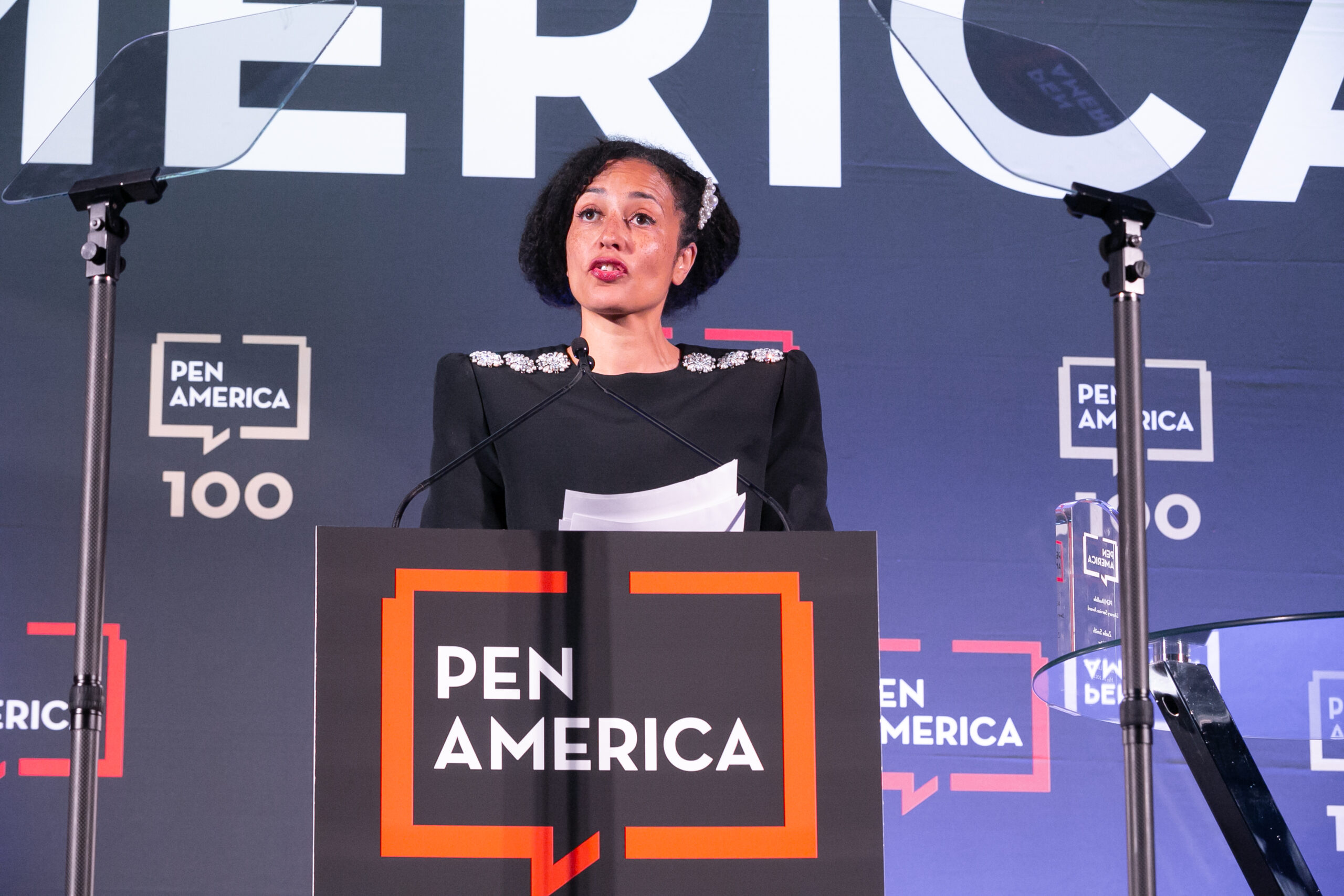 A woman with short dark hair speaks at a podium with a microphone during the PEN America Literary Gala. She holds papers and stands before a backdrop showing the PEN America logo.