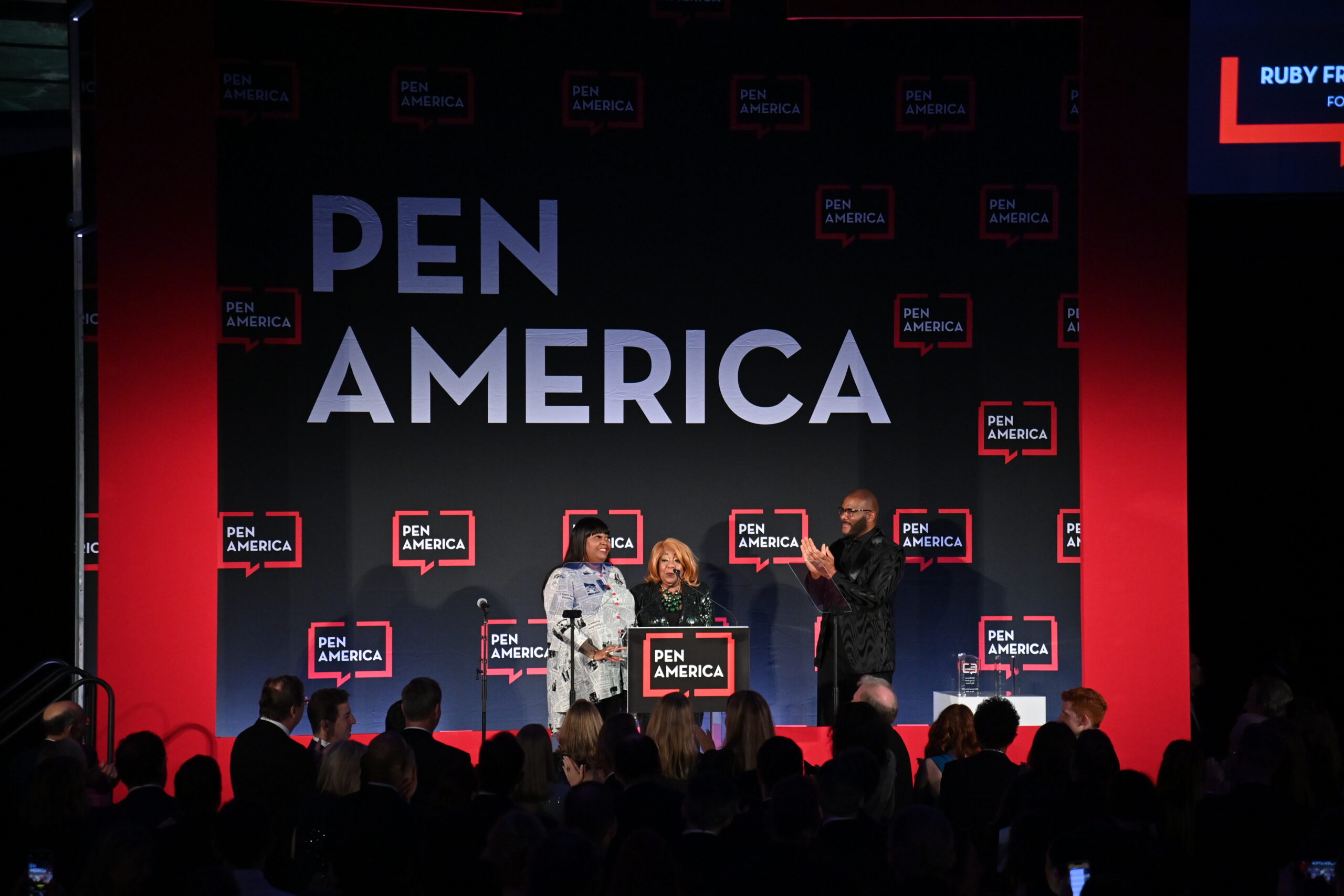 Three people stand on stage at a PEN America event, one at a podium labeled “PEN America,” with a large audience in front of them and a backdrop displaying the PEN America logo multiple times.