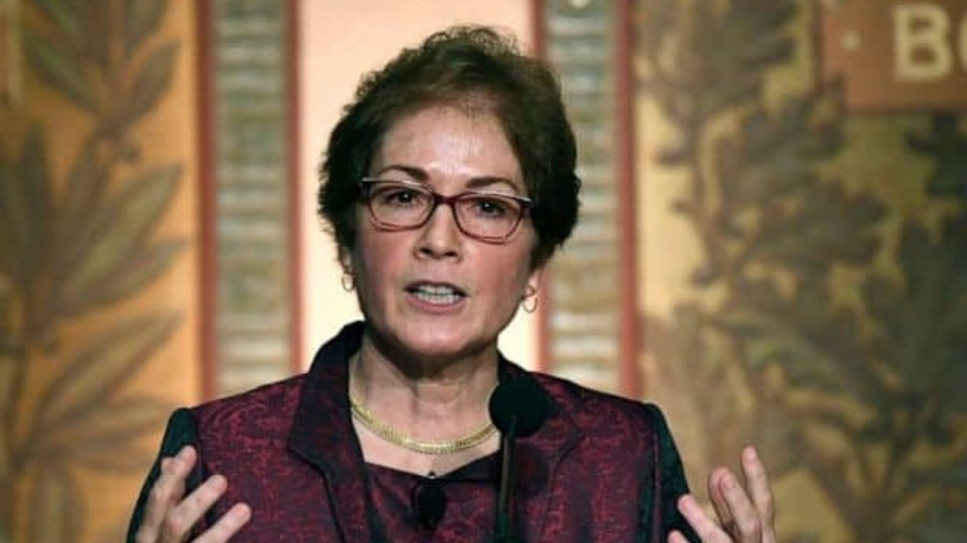 A woman wearing glasses and a maroon jacket speaks at a podium, gesturing with both hands. Ornate patterns and decorative leaves are visible on the wall behind her.