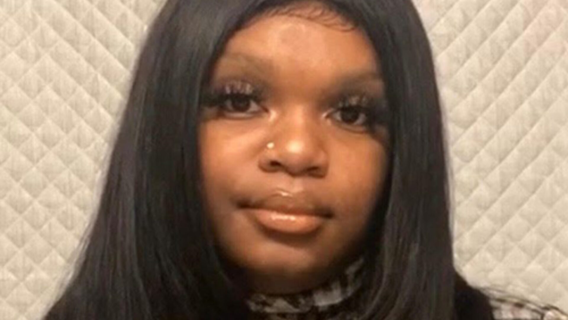 A woman with long straight black hair and a nose piercing is looking at the camera. She is wearing false eyelashes and a patterned top, with a quilted light gray background behind her.