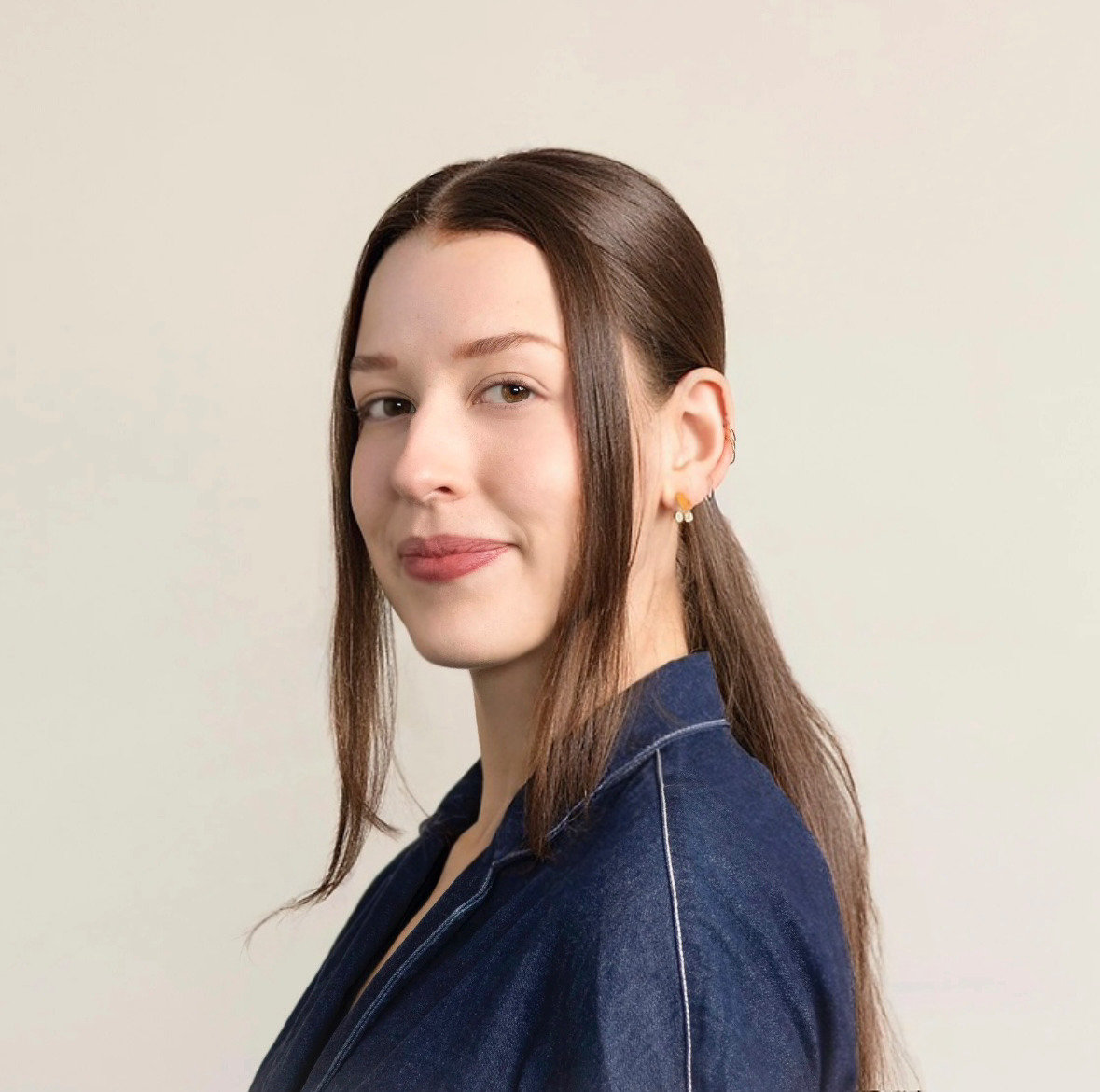 A woman with long, straight brown hair is smiling slightly at the camera. She is wearing a dark blue collared shirt and has multiple earrings. The background is a plain, light color.