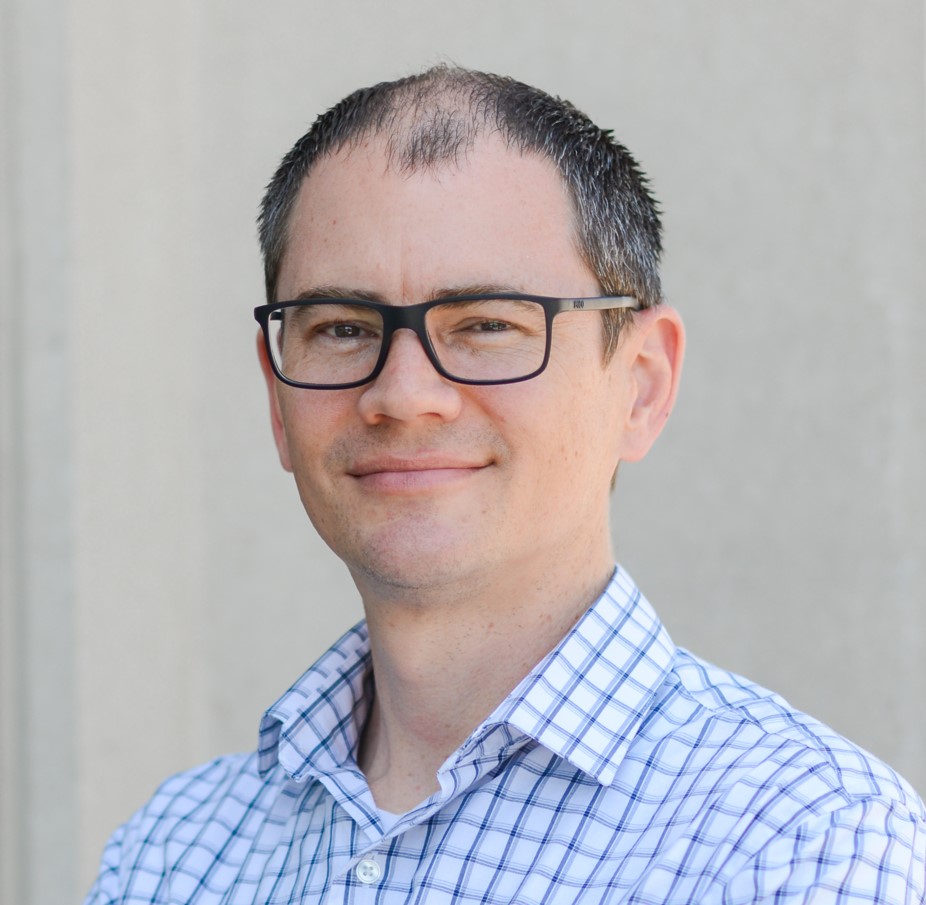 A man with short dark hair, glasses, and a checkered white and blue shirt stands smiling in front of a plain light-colored background.