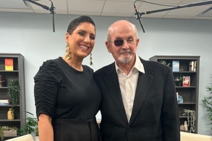 A woman in a black dress stands next to an older man in a black suit and sunglasses. They are both smiling and posing indoors with bookshelves and microphones in the background.