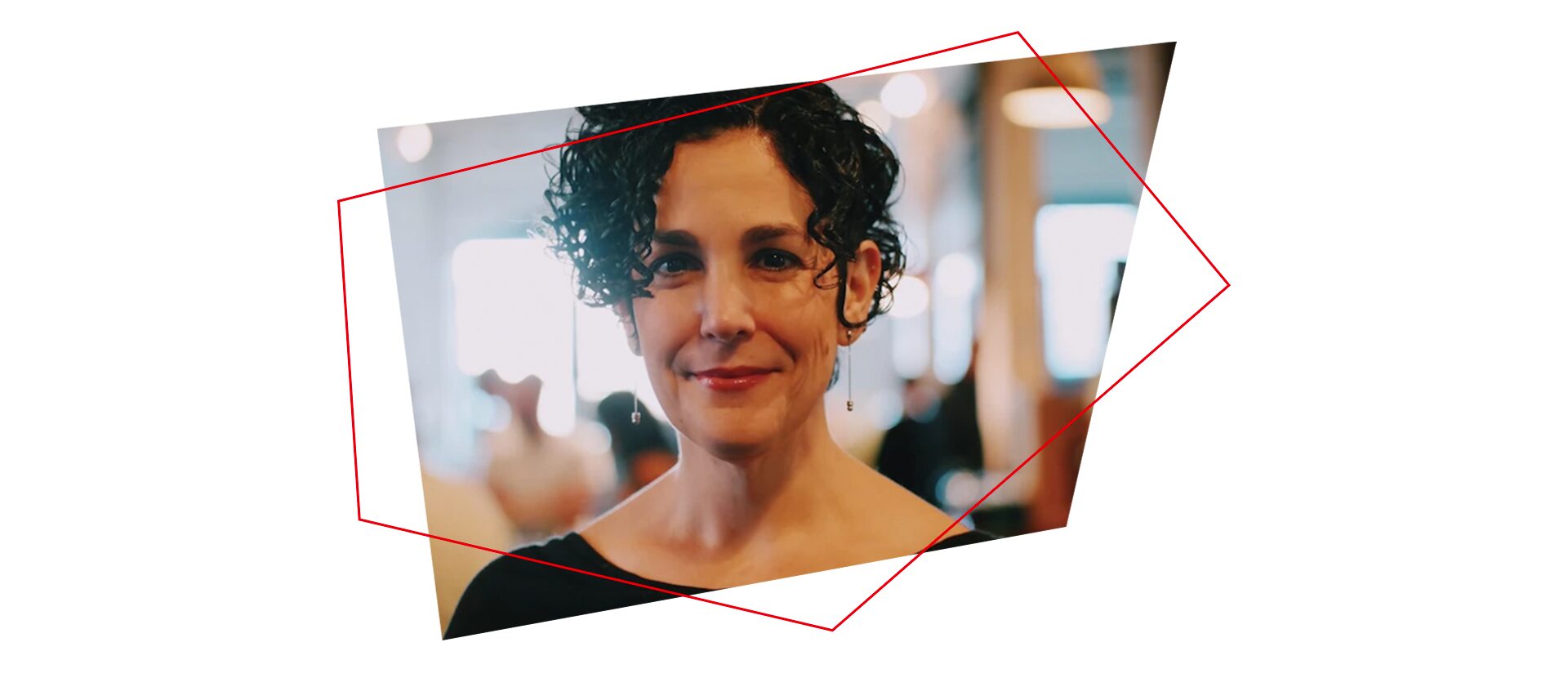 Yael Eisenstat, with short curly dark hair, wearing a black top and small earrings, smiles softly while standing indoors against a blurred background. Red geometric lines frame the image.