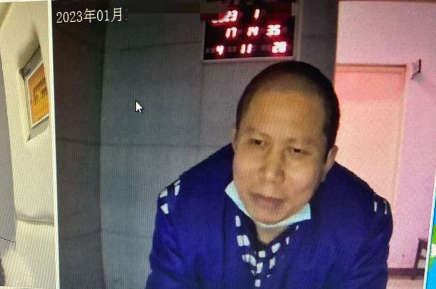 A man wearing a blue jacket and a partially lowered face mask leans forward indoors. A digital clock displaying red numbers and the text 2023年01月 are visible in the background.