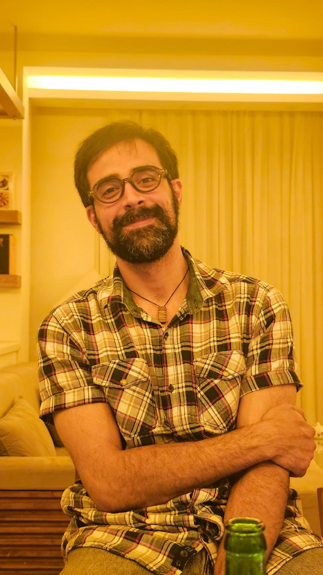 A bearded man wearing glasses and a plaid shirt sits indoors, smiling with his arms crossed. The room has warm yellow lighting, with a sofa and curtains in the background. A green bottle is visible in the foreground.