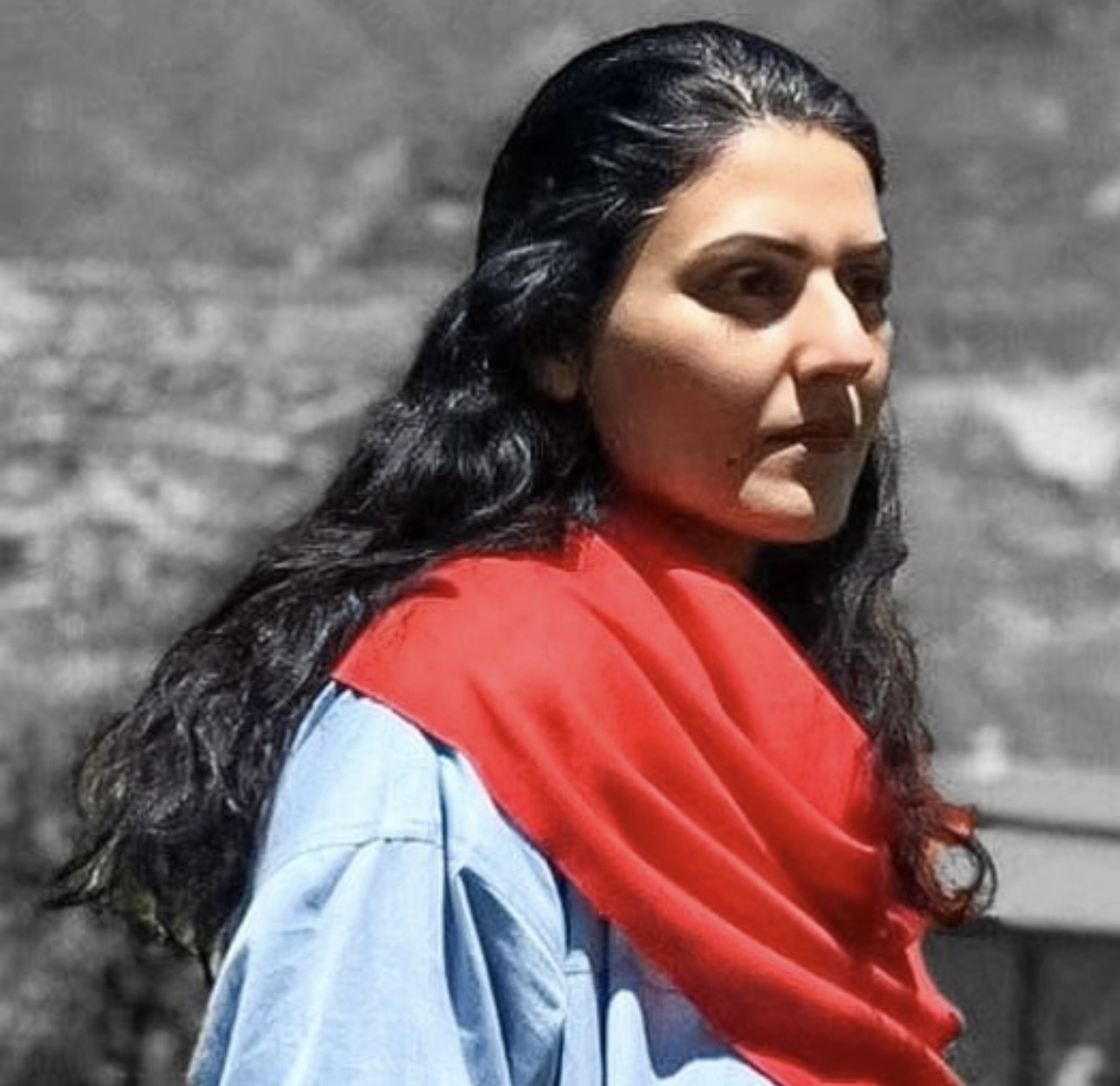 A woman with long dark hair wearing a red scarf over a light blue garment stands outdoors with a blurred, grayscale natural background.