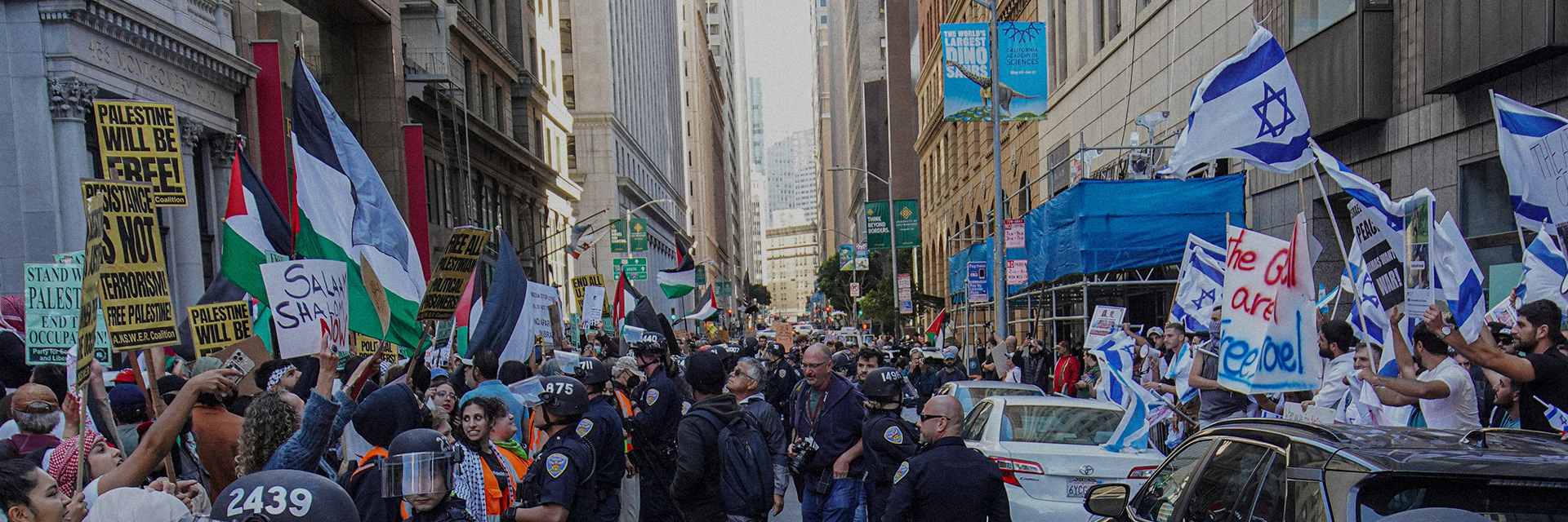 A busy city street features a large protest with two groups. One side holds Palestinian flags and signs, the other displays Israeli flags and banners. Police officers observe the scene, and people fill the sidewalks. Tall buildings line the street.