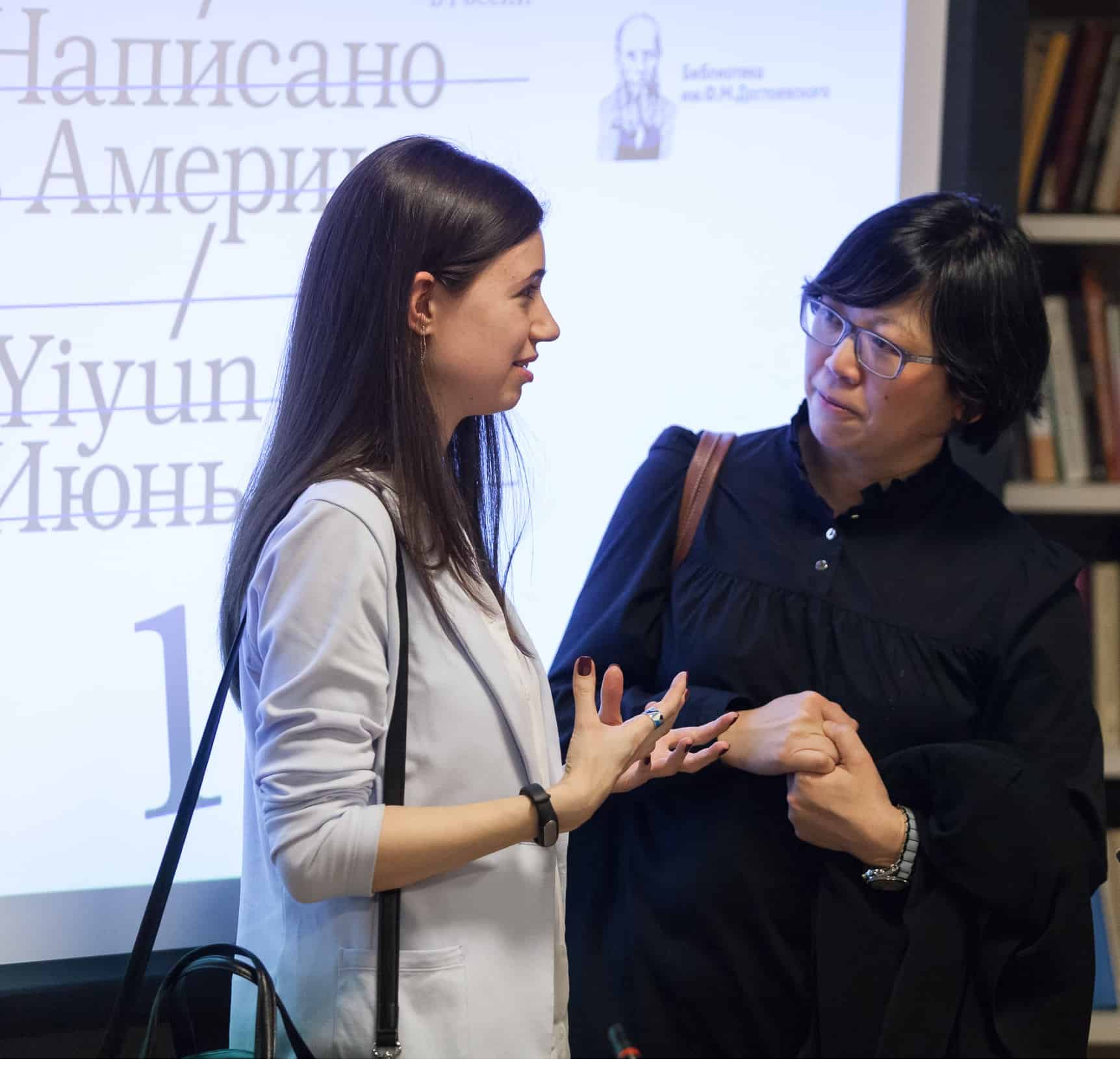 Two women stand and talk in front of a projected screen displaying text in Russian and English. One gestures with her hands, while the other listens attentively, holding her coat. Bookshelves are visible in the background.