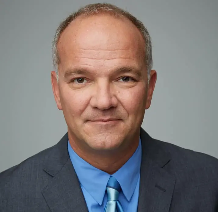 A middle-aged man with short light brown hair, wearing a blue dress shirt, blue tie, and dark suit jacket, poses against a plain gray background.