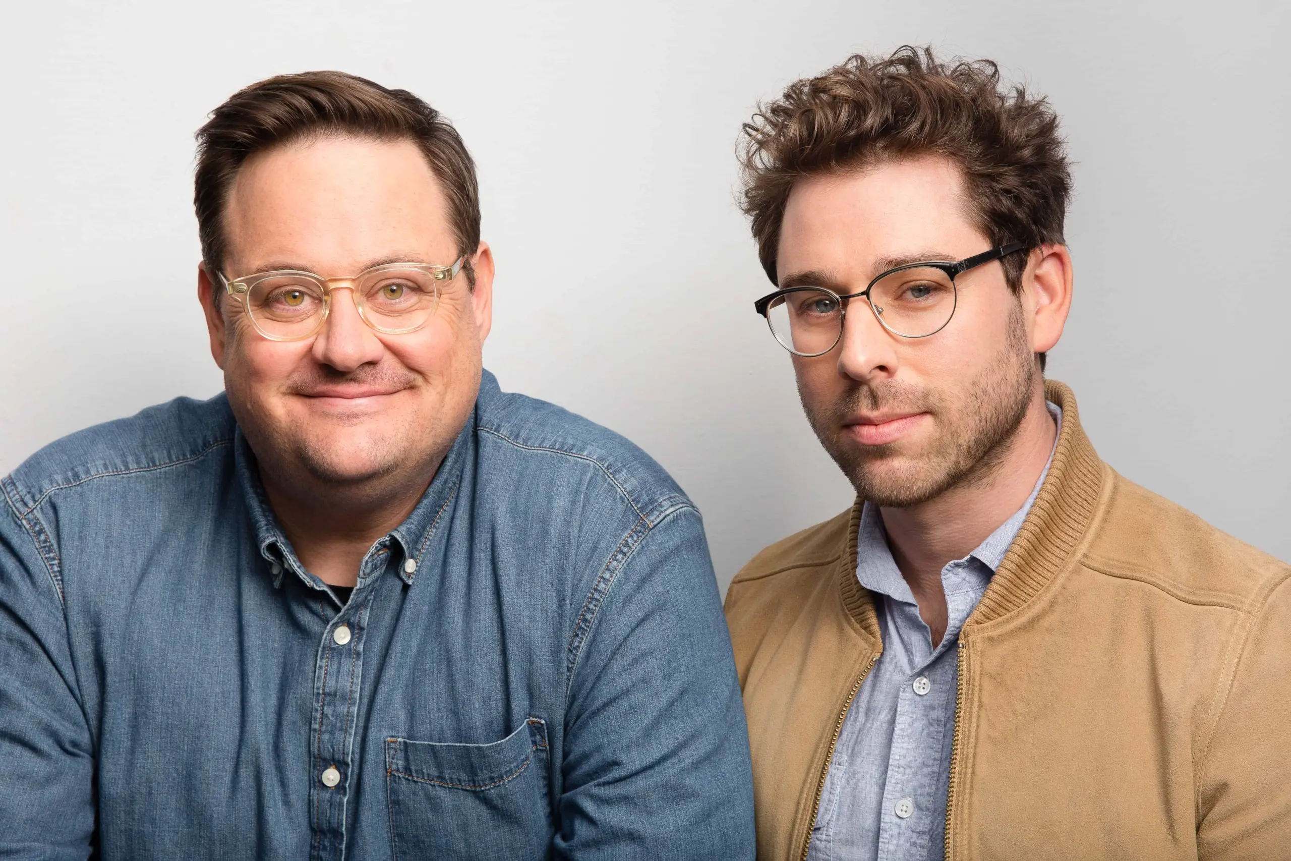 Two men with glasses pose in front of a plain light background. One wears a denim shirt and smiles, while the other wears a tan jacket over a blue button-down and has a neutral expression.