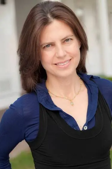 A woman with straight brown hair and fair skin smiles at the camera. She is wearing a blue collared shirt under a black sleeveless top and a gold necklace. The background is softly blurred.