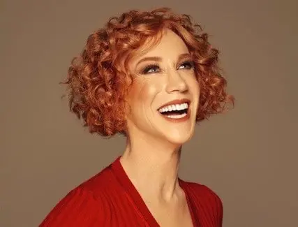 A woman with short, curly red hair smiles brightly while looking upward. She is wearing a red top and has a warm, neutral background behind her.