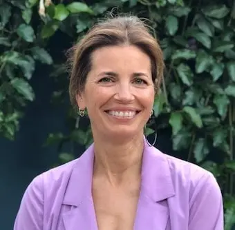 A woman with light brown hair in an updo, wearing a light purple blazer and hoop earrings, smiles while standing in front of green leafy plants—radiating the confidence of a 2019 LitFest Gala honoree.