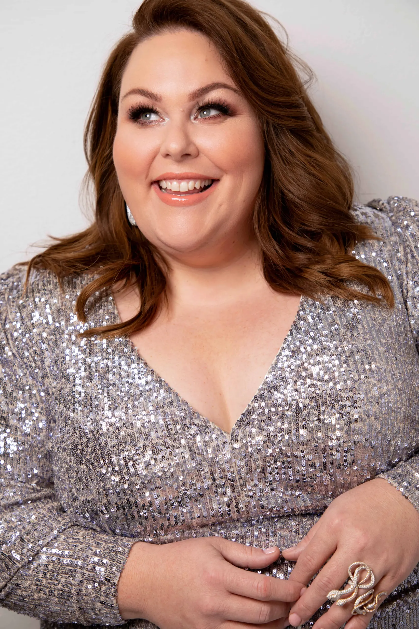 A smiling woman with wavy brown hair, the 2019 LitFest Gala host, wears a sparkly silver sequin dress with puffed shoulders. She has natural makeup and looks slightly upward, appearing cheerful and confident.