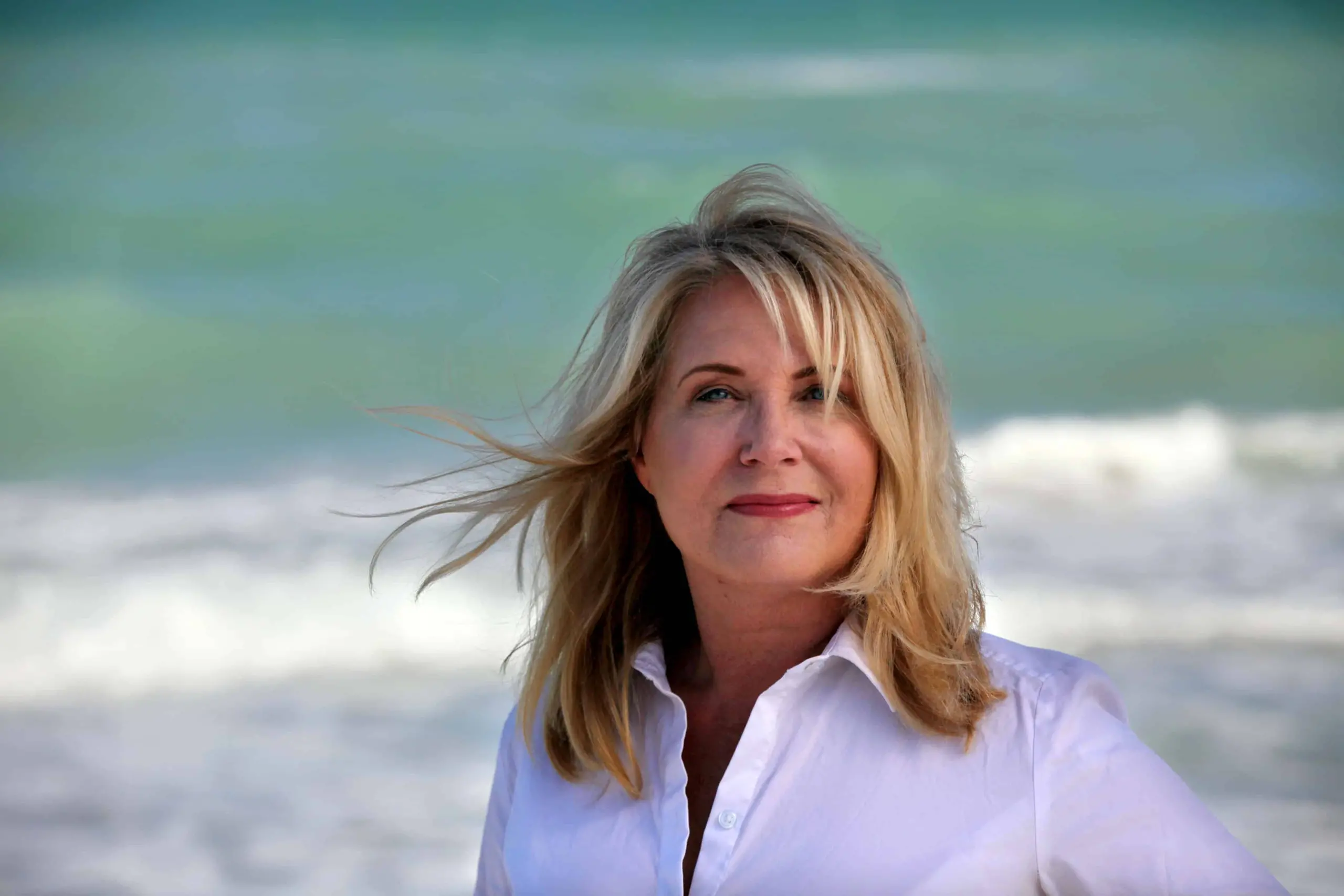 A woman with blonde hair, the 2019 LitFest Gala host, stands in front of the ocean, her white shirt bright against the waves and blue water as her hair gently blows in the wind.