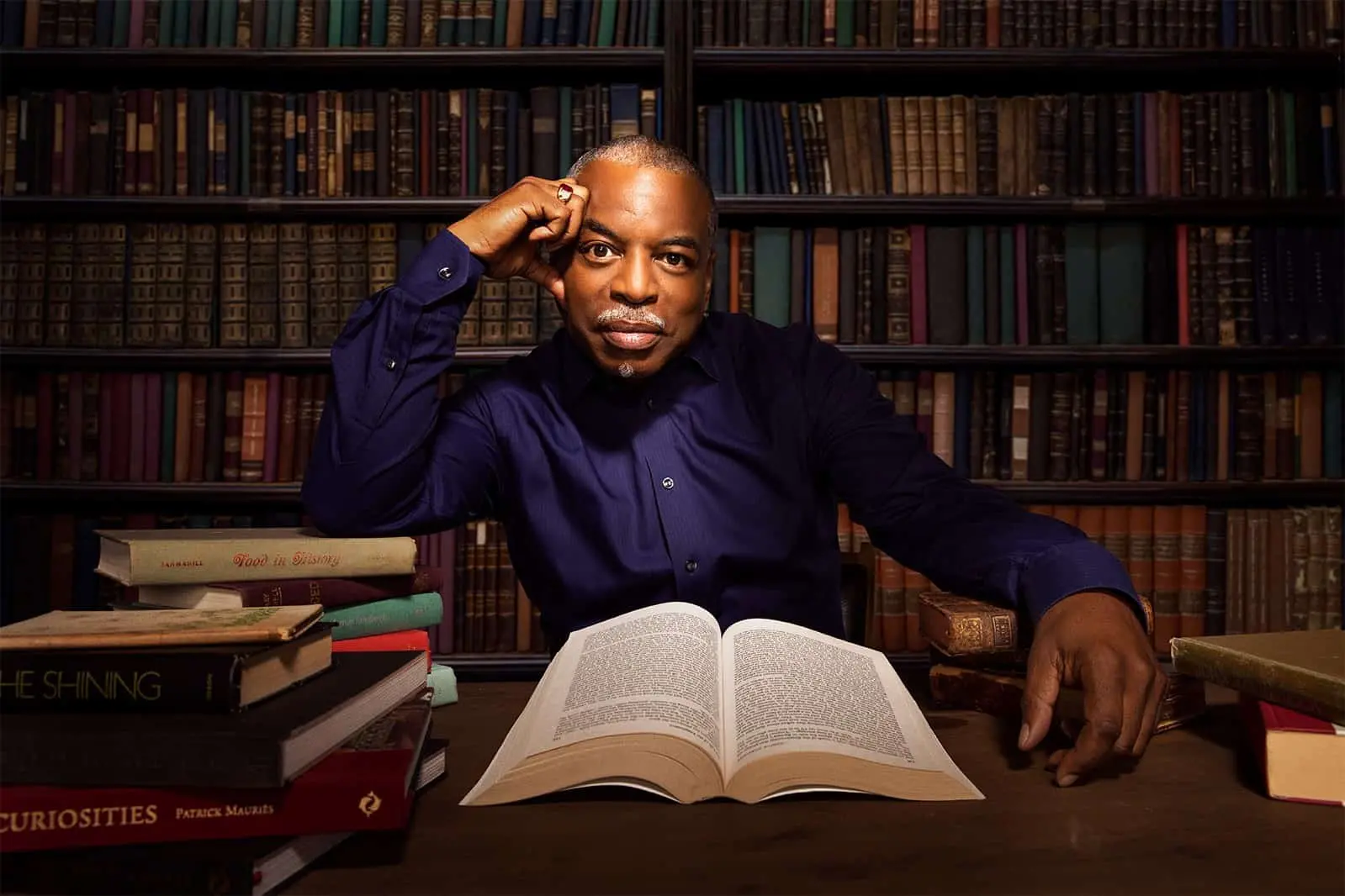A 2019 LitFest Gala honoree in a dark blue shirt sits at a desk with an open book in front of him, resting his head on his hand, surrounded by stacks of books and a wall of bookshelves in the background.