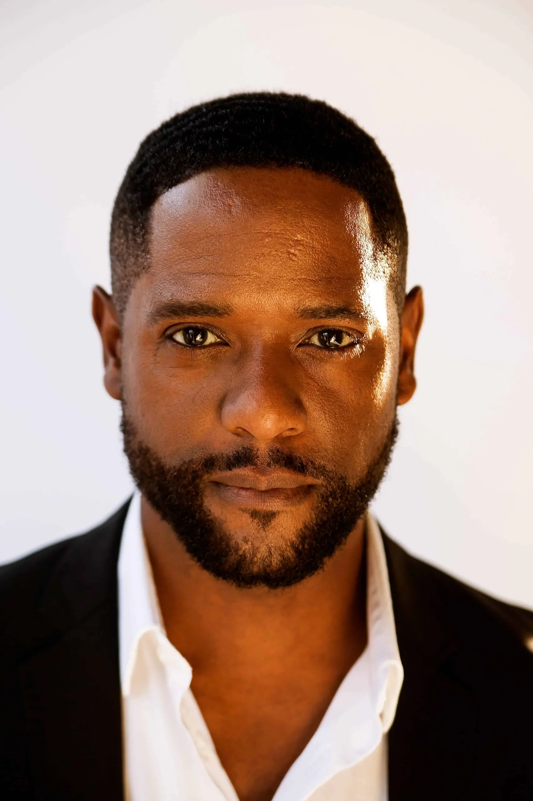 A man with short black hair and a trimmed beard, dressed in a white shirt and black jacket, looks directly at the camera with a neutral expression against a plain light background at the 2019 LitFest Gala, reminiscent of Blair Underwood's poised style.
