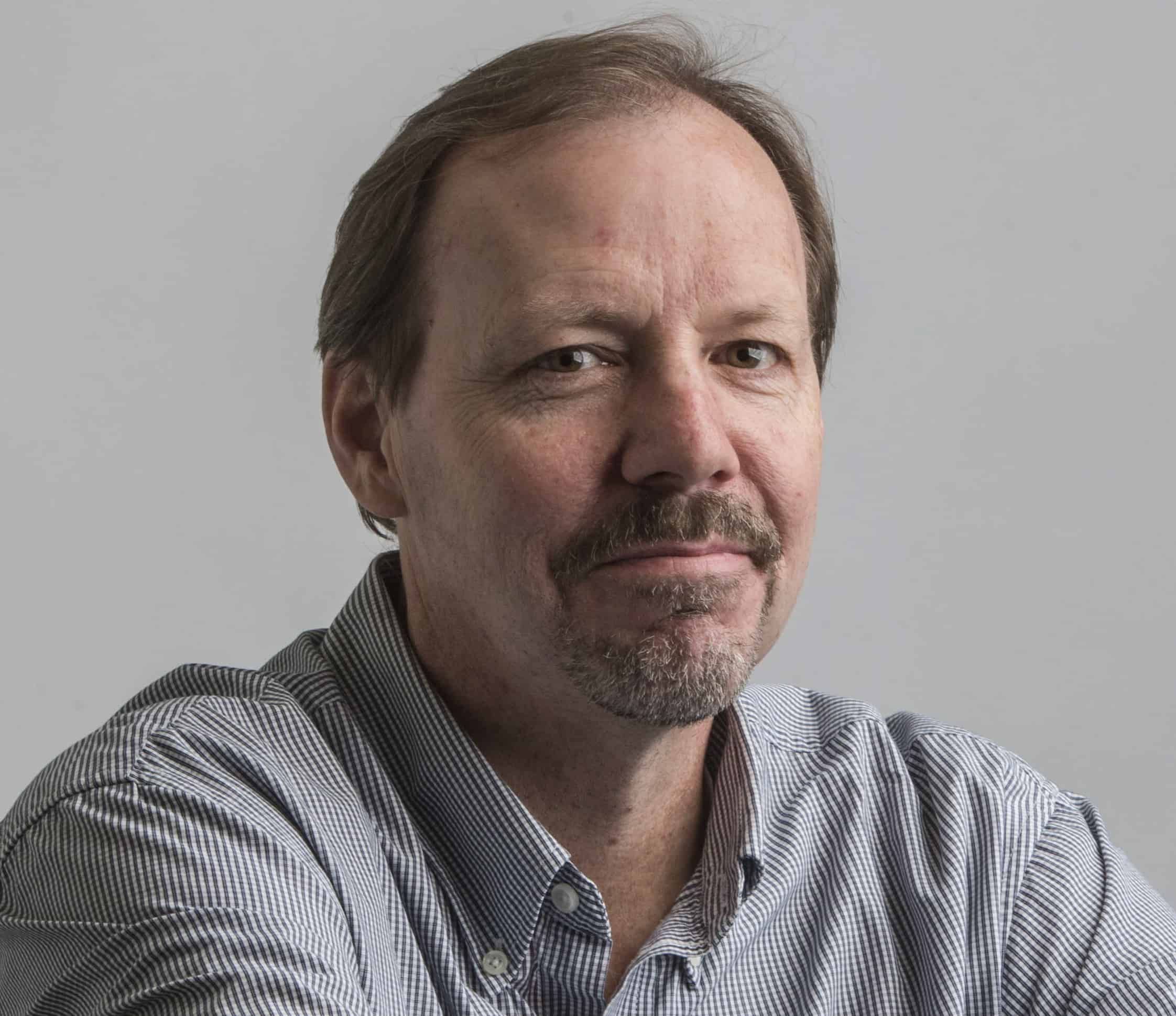 A middle-aged man with short brown hair, a mustache, and a goatee—resembling 2019 LitFest Gala guest Ken Armstrong—wears a striped button-up shirt and looks at the camera with a neutral expression against a plain gray background.