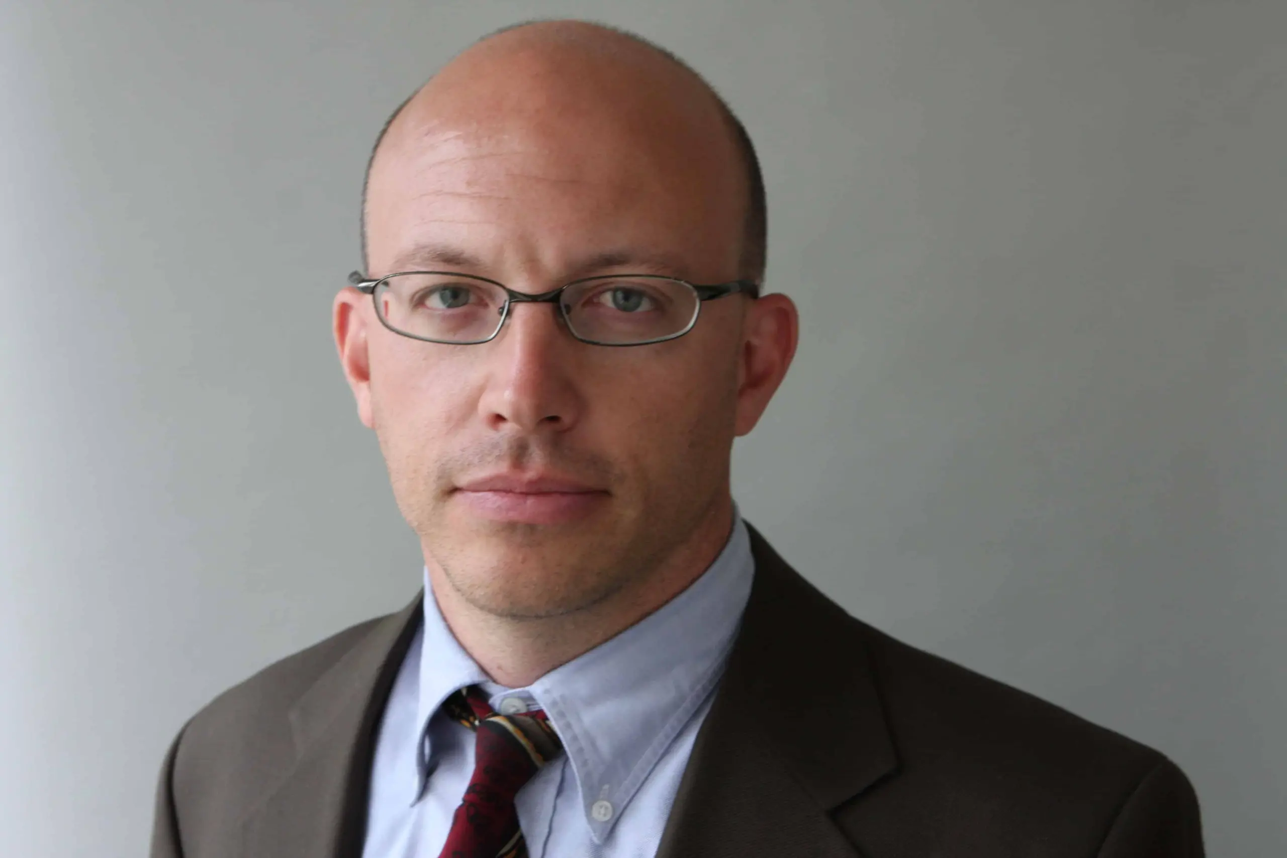 A man with glasses, a bald head, and light skin wearing a suit, dress shirt, and striped tie looks directly at the camera against a plain gray background—reminiscent of 2019 LitFest Gala guest T. Christian Miller.