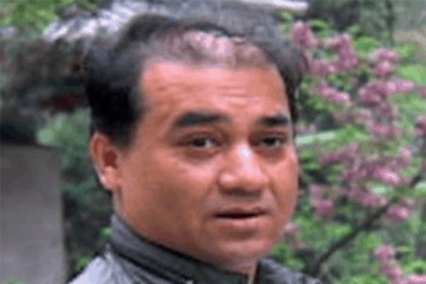 A man with short black hair and a slight receding hairline looks toward the camera. He is outdoors, with greenery and purple flowers visible in the blurry background.