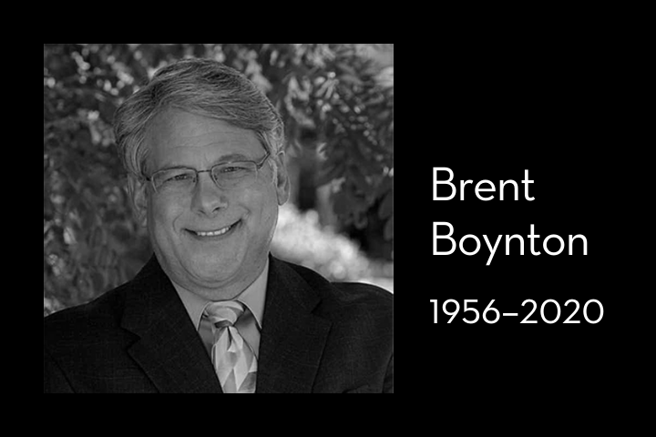 Black and white portrait of a smiling man in a suit and tie, with gray hair and glasses. Text beside him reads: Brent Boynton 1956–2020. Background includes tree foliage.