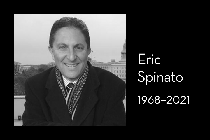 Black-and-white photo of a smiling man in a coat and patterned scarf outdoors, with buildings in the background. Text beside reads: Eric Spinato 1968–2021.