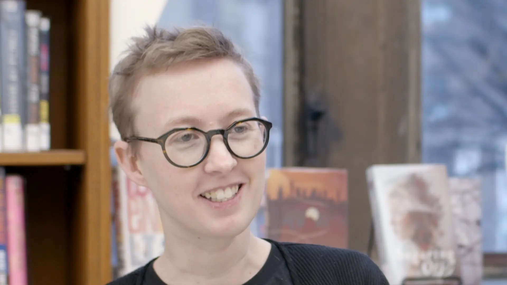 A person with short, light hair and round glasses smiles while sitting indoors, with bookshelves and books visible in the background.