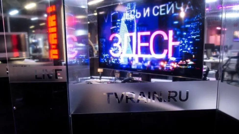 A glass-walled TV studio with a monitor displaying Russian text and colorful city lights, and TVRAIN.RU etched on the glass wall in front of the studio.