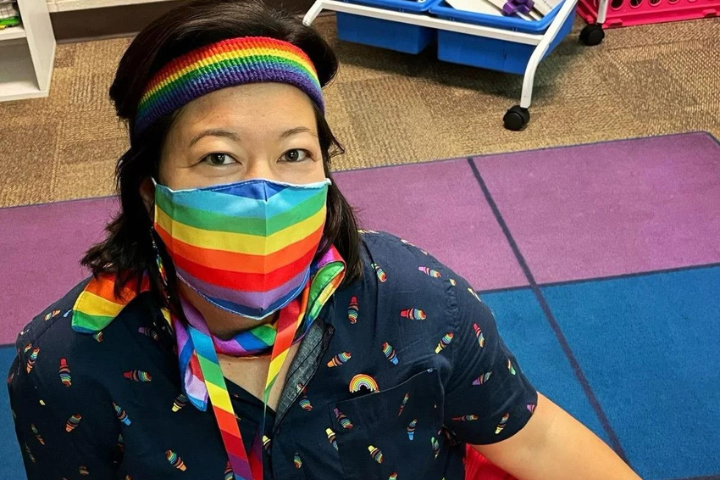 A person wearing a rainbow headband, rainbow face mask, and rainbow scarf sits on a colorful carpet. Their shirt also has rainbow designs, and classroom furniture is visible in the background.