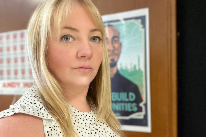 A woman with blonde hair and a white polka-dot top stands indoors near a wooden wall with posters, including one featuring a mans illustrated portrait in the background.