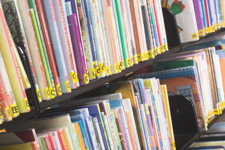 Rows of colorful childrens books with yellow spine labels are neatly arranged on library shelves, viewed from an angle. Some book covers and titles are partially visible.