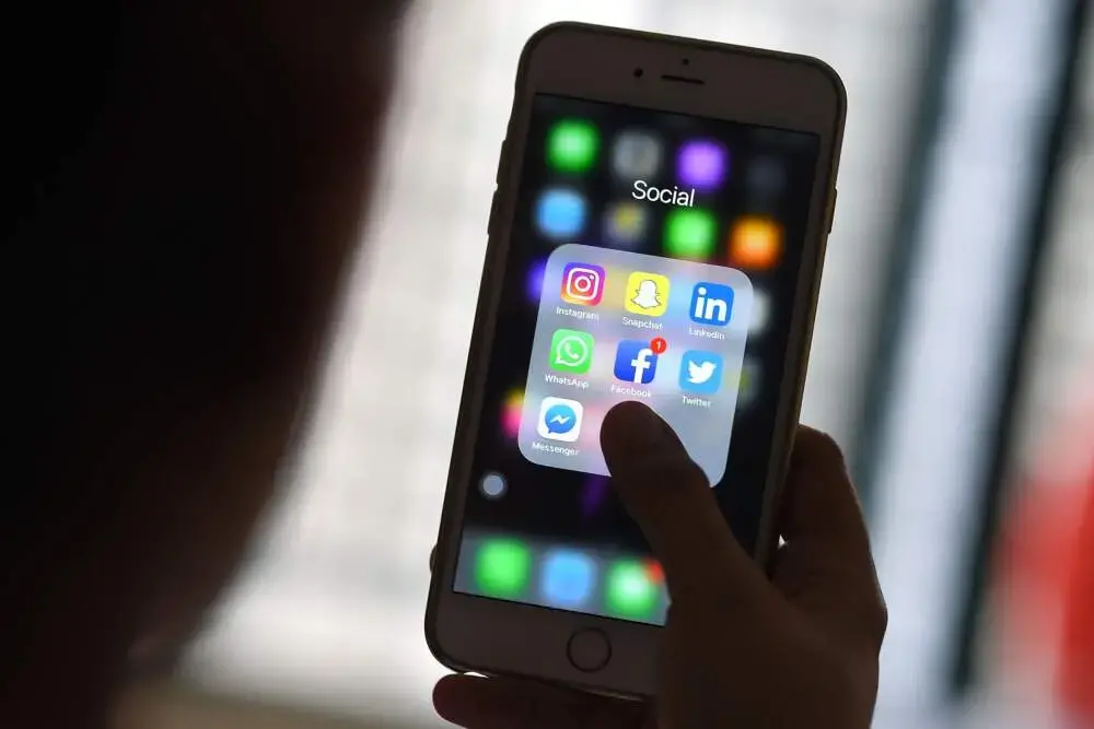 A person holds a smartphone and taps a folder labeled “Social,” which contains app icons for Instagram, Snapchat, LinkedIn, WhatsApp, Facebook, Twitter, and Messenger. The background is blurred.