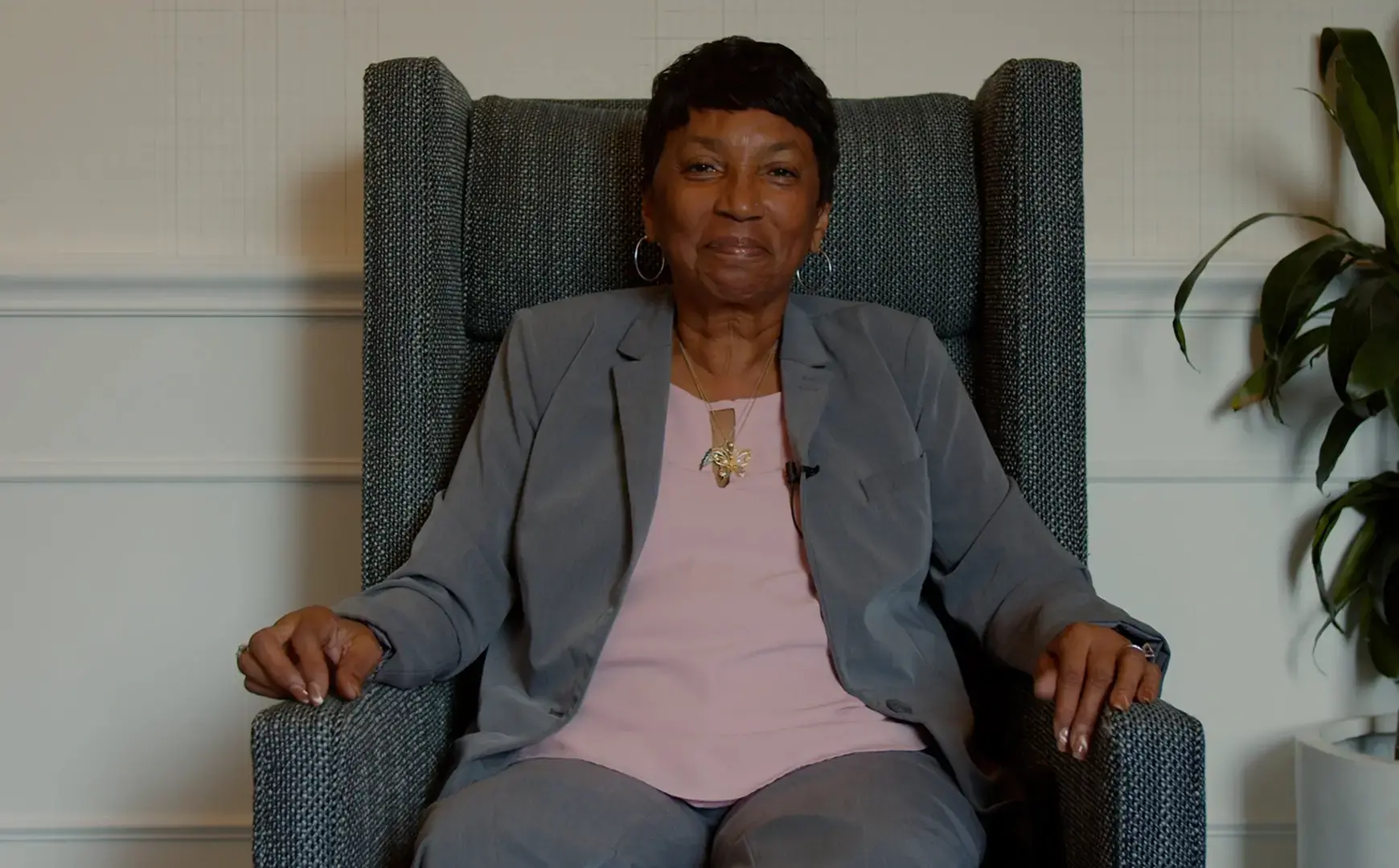 A woman wearing a gray suit and a light pink top sits in a large, high-backed chair, smiling slightly at the camera. She has short hair and is wearing hoop earrings and a butterfly necklace. A plant is visible to the right.