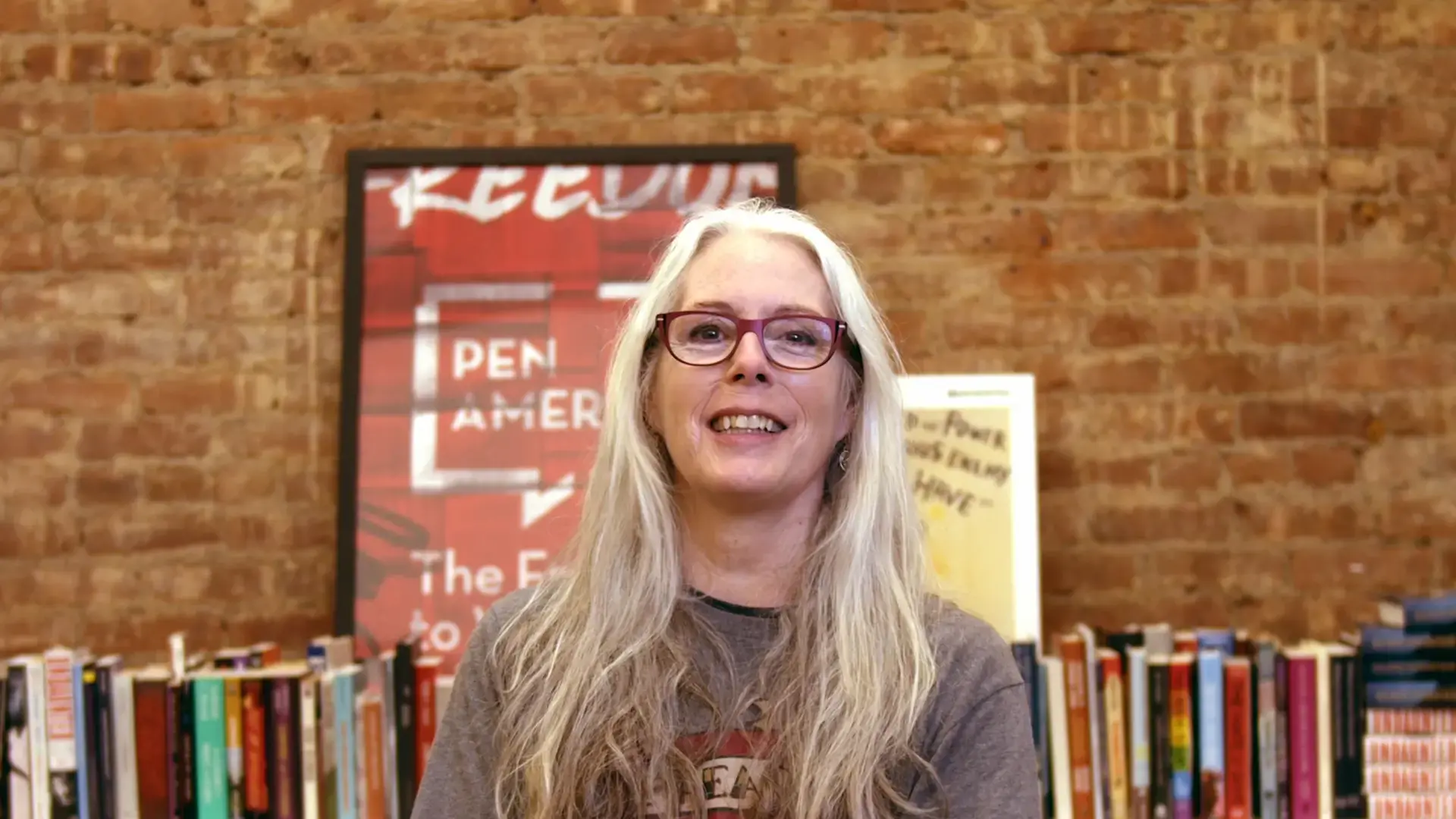 A woman with long gray hair and glasses smiles while sitting in front of a bookshelf and a brick wall. A framed poster and several books are visible in the background.