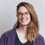 A person with long, wavy, light brown hair wearing glasses, a nose ring, a black shirt, a patterned blazer, and a necklace—smiling in front of a plain light-gray background—represents the spirit of PEN’s NYC Free Expression Advocacy Institute.
