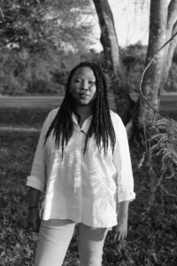 A woman with long braids stands outdoors before trees, wearing a light-colored button-up shirt and pants. Dappled sunlight creates shadows on her shirt in this black and white portrait of a PEN Bellwether Prize nominee.