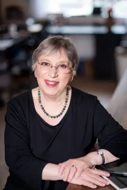 An older woman with short gray hair, glasses, and a colorful beaded necklace smiles while sitting at a desk, wearing a black top and watch, with her hands resting on the table. The background is softly blurred.