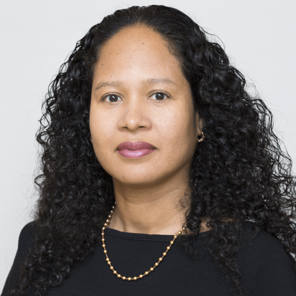 A woman with long, curly black hair and medium skin tone, wearing a black top, gold necklace, and small hoop earrings, poses against a plain light background—echoing the spirit of NYC Free Expression Advocacy Institute PEN.