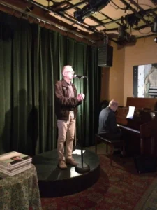 A man stands on a small round stage speaking into a microphone, while another man plays a piano nearby. They are indoors, in front of dark green curtains, with a stack of books visible on a table.